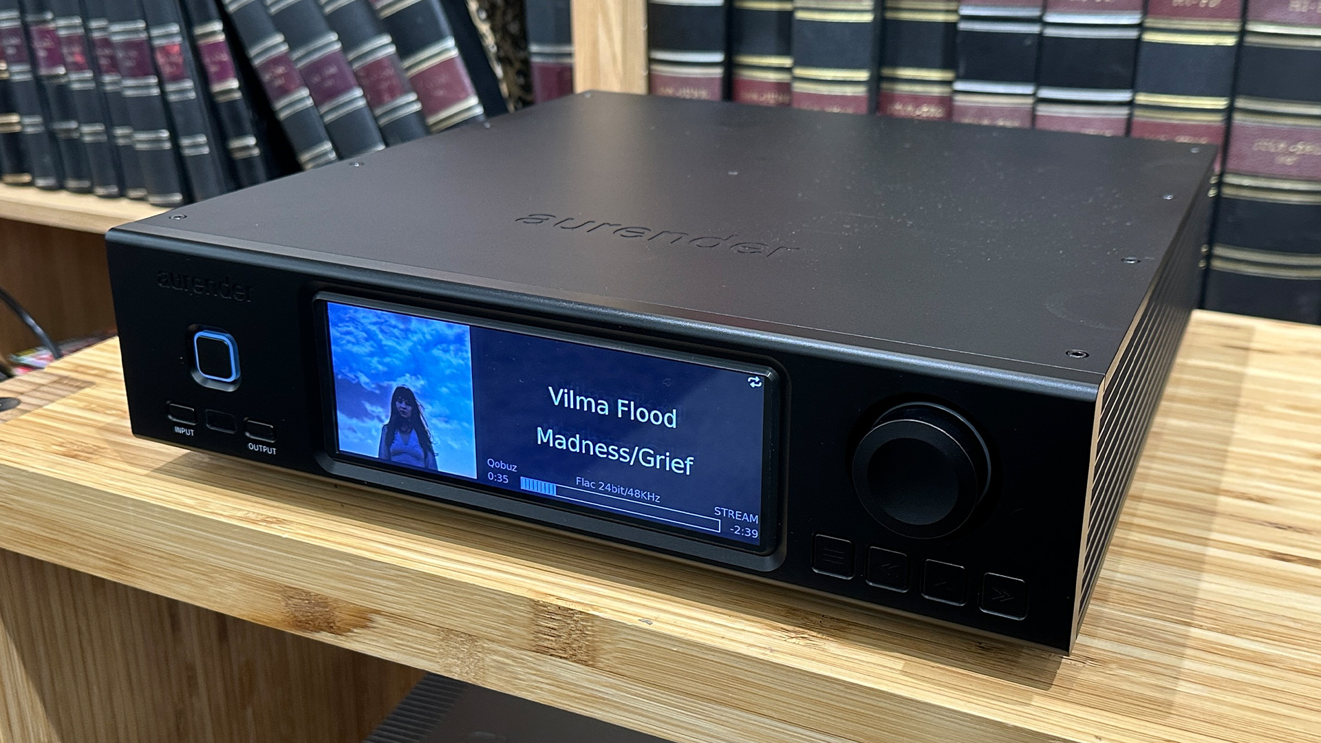 Aurender A1000 music streamer on wooden hi-fi rack in front of bookcase, slight angle showing Vilma Flood on display