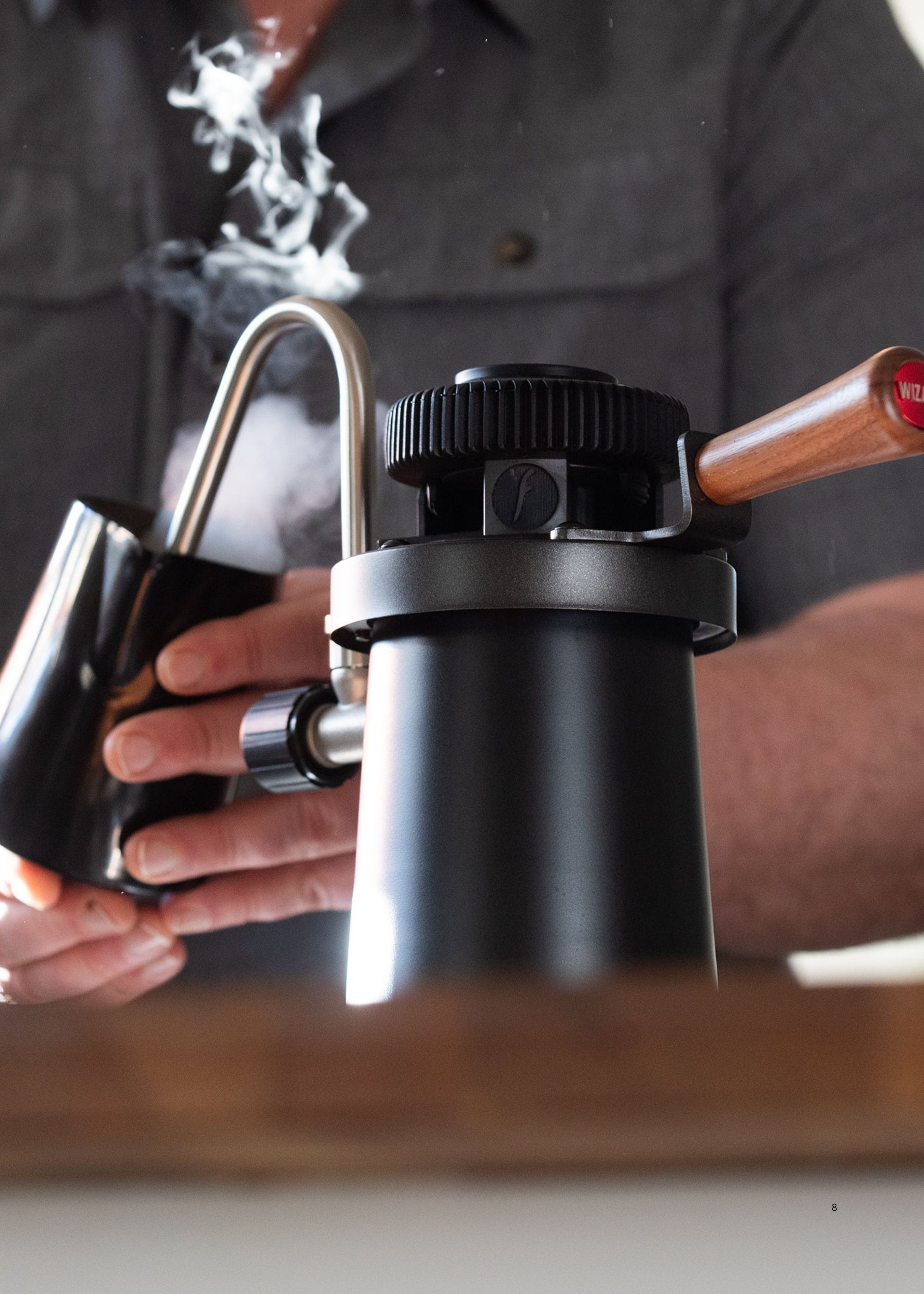 Image of a person in a gray shirt holding a black milk steaming jug under a silver steaming wand steaming milk from a black Flair Wizard Stovetop Milk Steamer.