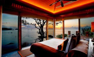 Habita hotel room with high wooden ceilings, rust bedding and floor to ceiling windows overlooking sunset on sea.
