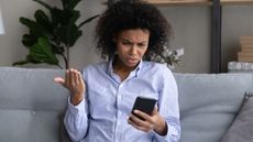 A woman looks at her phone with an alarmed look on her face while sitting on her sofa.