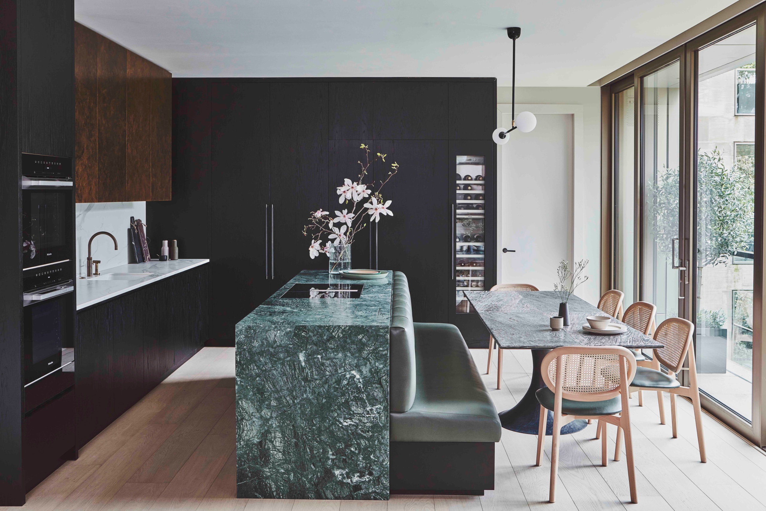 Green stone kitchen island with an upholstered banquette built into its dining-facing side, beside a rectangular pedestal table and wood-framed cane chairs overlooking full-height windows.