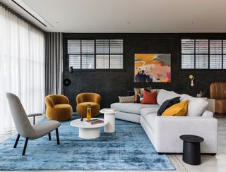Living-dining space with white angled sofa, mustard and grey armchairs, blue rug and black walls