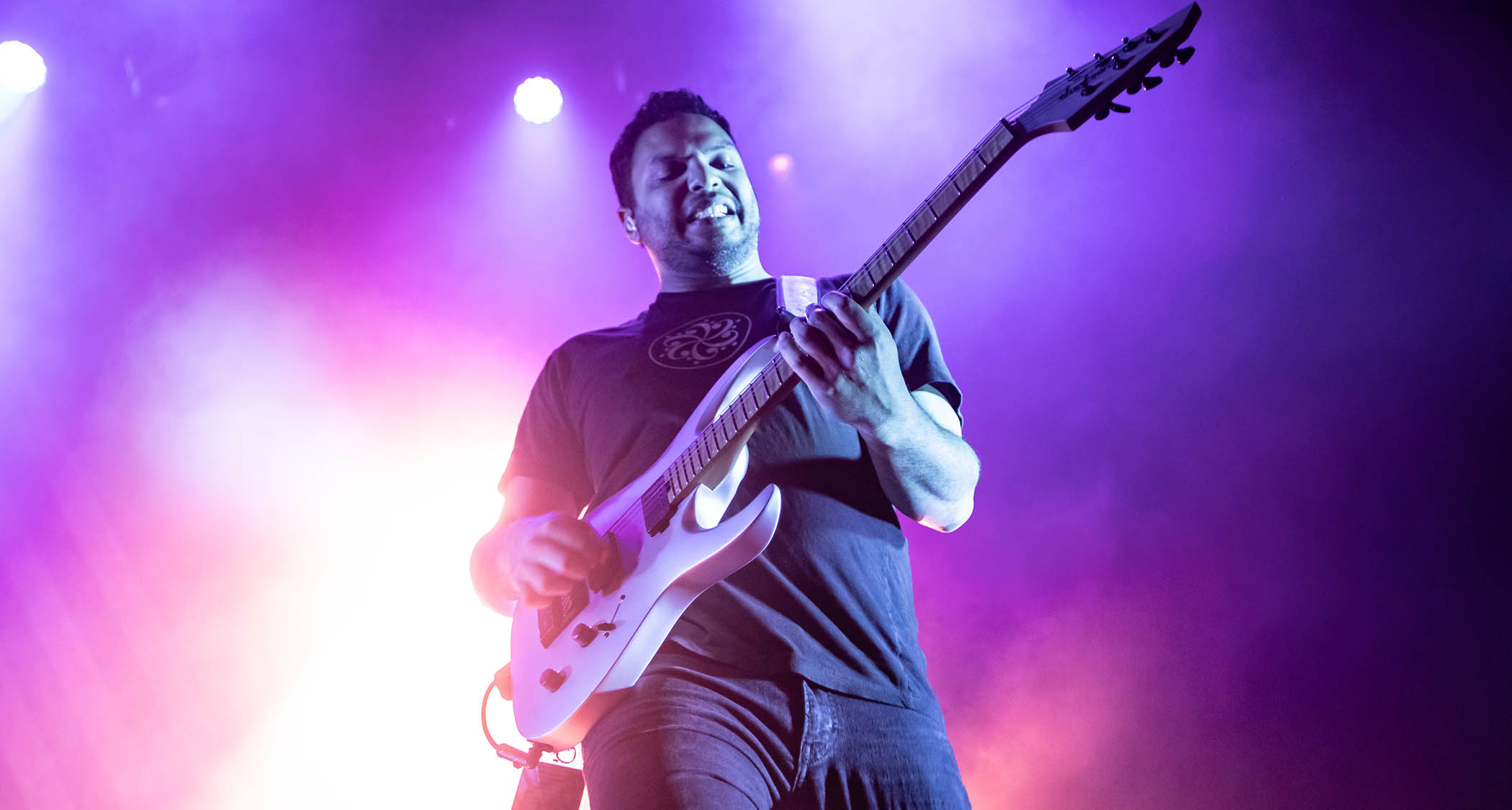 Misha Mansoor digs in on his signature Jackson Juggernaut as he play live with Periphery. The stage is lit in purple and white.