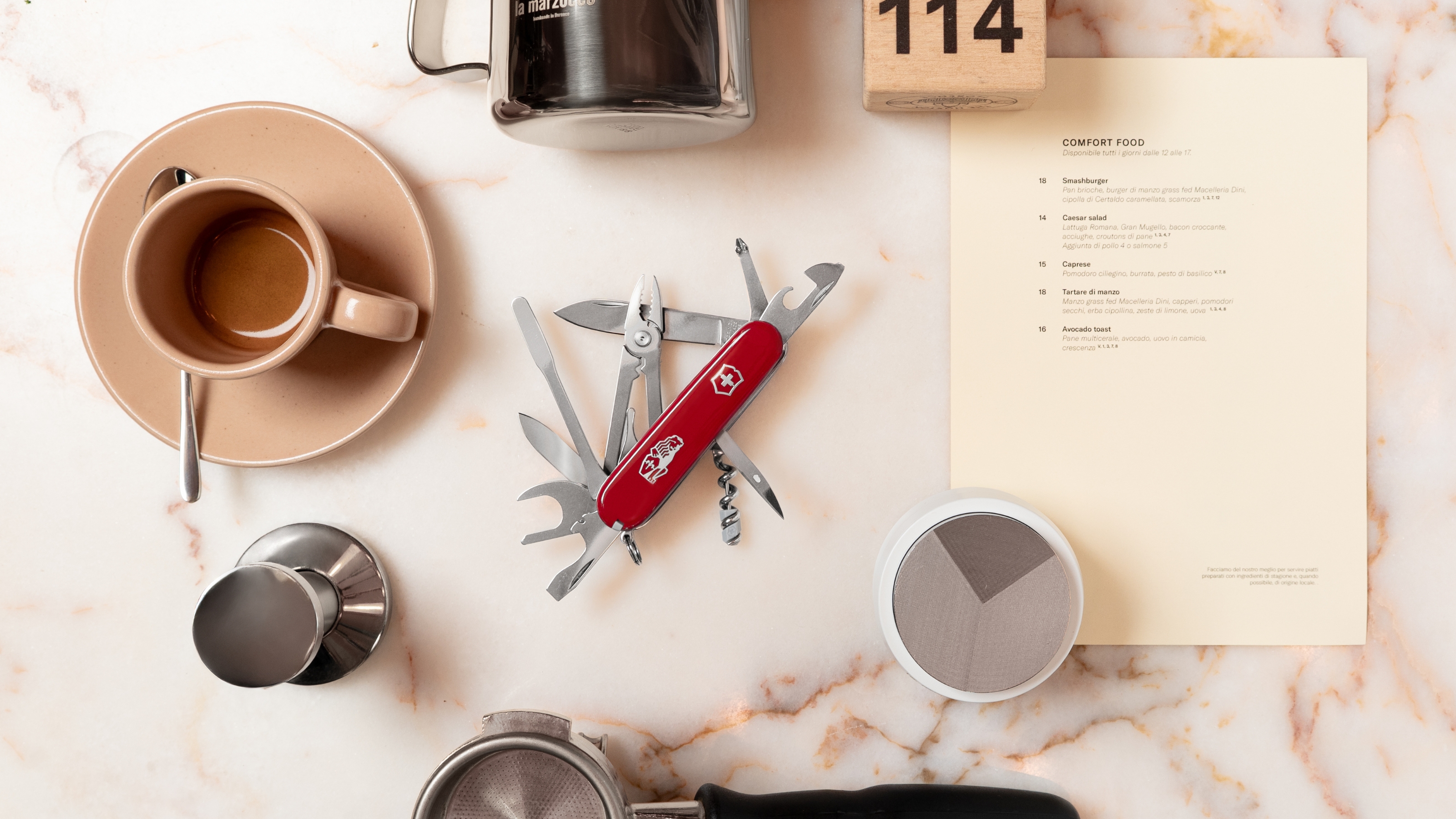 Image of a pocket knife style coffee tool with all of the gadgets open, sitting on a white marble countertop. There are several coffee accessories in a circle around it.