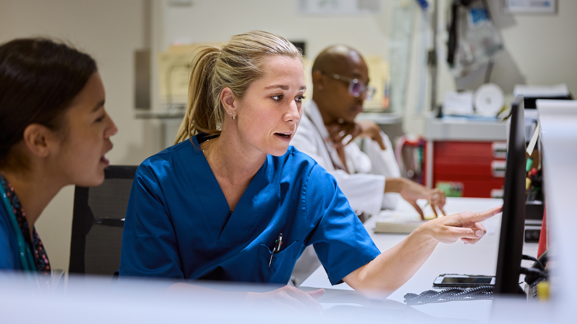 Two nurses wearing blue scrubs sit in front of a computer in a hospital