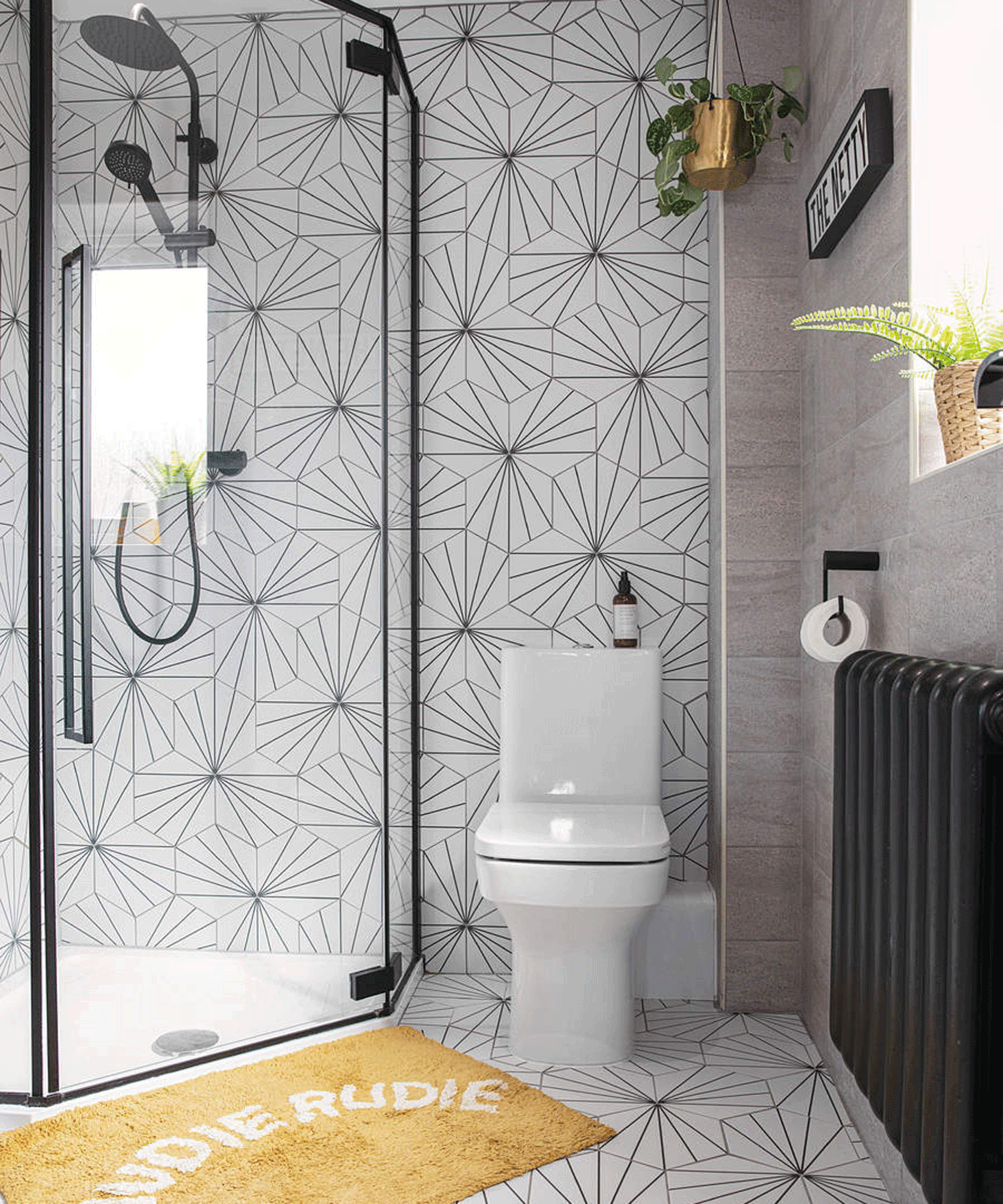 A contemporary bathroom with dual shower fixture, white geometric wall tile decor and slogan 'Nudie Rudie' yellow bath mat