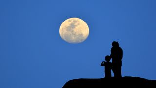 Silhouette of mother and child watching the moon against the blue night sky.