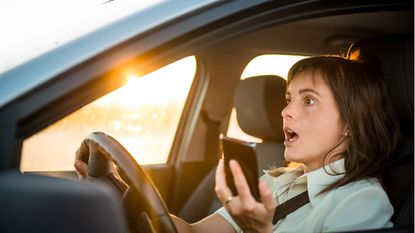 A woman holding her phone while driving looks alarmed.