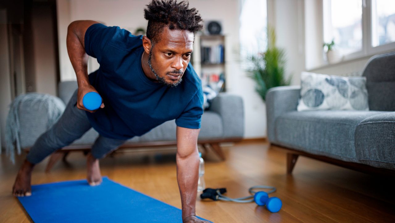 Man exercising at home, wearing blue T-shirt and gray exercise pants. He is in a high plank position, lifting a dumbbell to his torso 