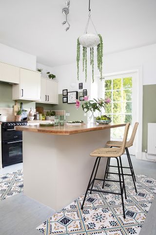 Kitchen with cream units, wood work surface, patterned floor tiles, green painted walls, rattan bar stools and plant in hanging planter