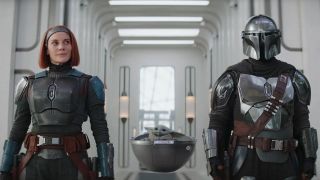 Still from the Star Wars TV Show, The Mandalorian. From left to right, we have Bo-Katan (shhoulder-length red hair mandalorian wearing blue armor), Grogu (small tiny green creature sitting in a floating 'pram'), and the Mandalorian himself (wearing full armor and helmet).