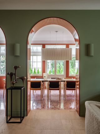 Image of a green living room with arched doorways that lead into an orange-themed dining room. The floors of the dining room are orange terrazzo. There are black and wood dining chairs around the dining table and a white paper lantern overhead.