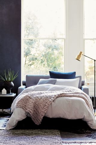 comfy sofa bed against window with pink and striped bedding, on top of a purple rug and next to a gold floor lamp