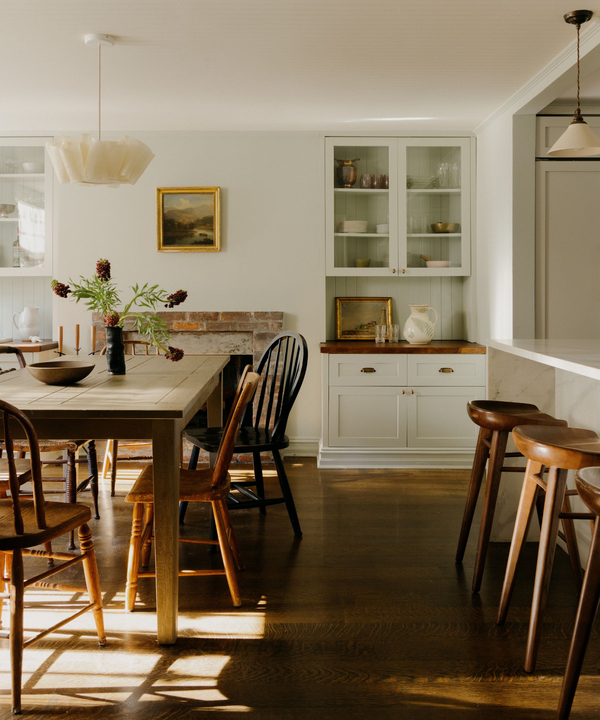 A warm, sunlit dining area featuring a wooden harvest table with mismatched vintage chairs. In the background, there is a brick fireplace and white built-in cabinetry with glass doors and wood countertops.
