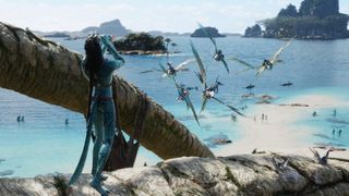 Still from the movie Avatar: The Way of Water (2022). Here we see a Na'vi (tall, blue skinned humanoids with lond dark hair) standing on a beach blowing into a conch shell. In the background you can see a number of winged-flying creatures and their Na'vi riders.