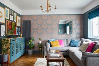 Living room with Art Deco-style feature wallpaper on one wall, teal paint on the others; parquet wood floor, grey L-shaped sofa, wooden coffee table and ornate blue and wood cabinets