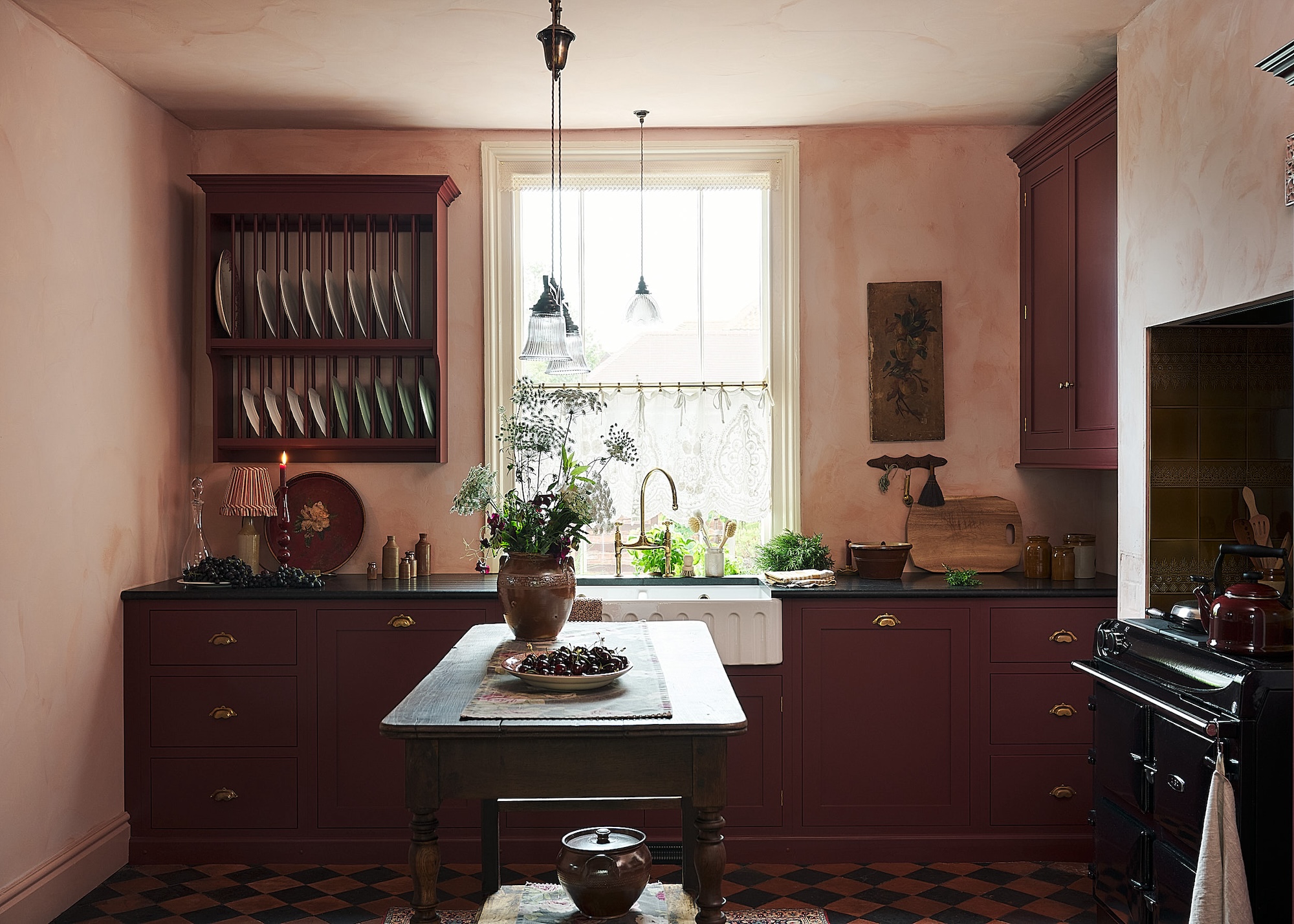 A pastel pink and deep maroon kitchen with a plate rack on the wall as well as a mini island placed at the centre of the kitchen