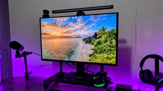 A computer monitor displays a vibrant beach sunrise scene. Surrounding it are a microphone, headphones, and colorful LED lights, creating a cozy tech setup.