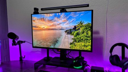 A computer monitor displays a vibrant beach sunrise scene. Surrounding it are a microphone, headphones, and colorful LED lights, creating a cozy tech setup.