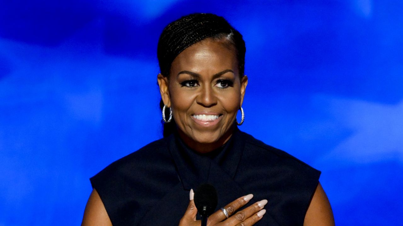 michelle obama puts her hand on her heart as she stands at a podium wearing a navy dress