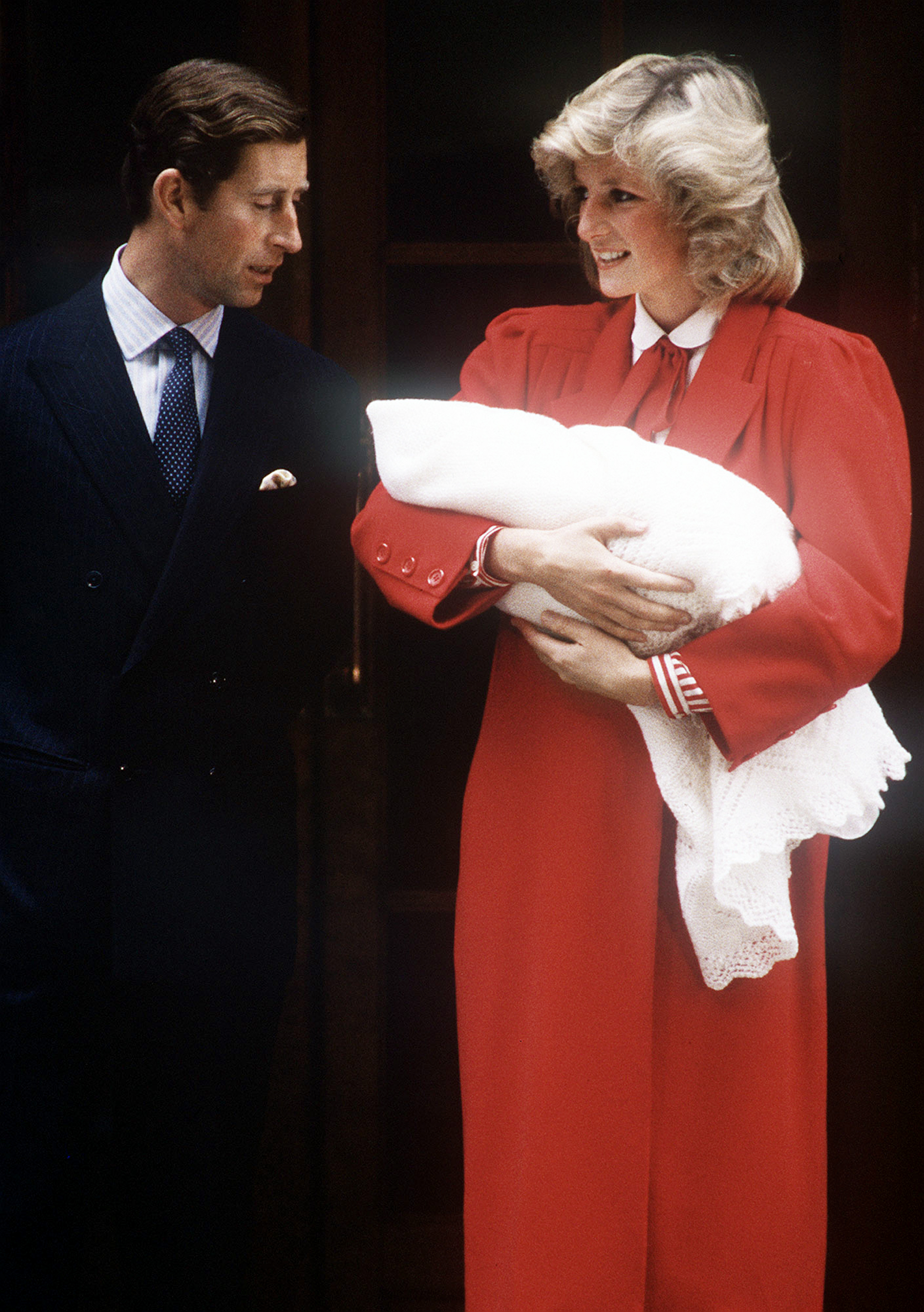 Princess Diana wearing a red coat holding Prince Harry in a blanket outside the hospital with Prince Charles