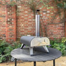 Drew&Cole pizza oven on a black garden table in front of a brick exterior wall 