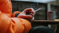 A man sits outside and holds a slim menthol cigarette between his fingers