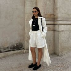 Influencer @deborabrosa wears white linen shorts with a black top, a white trench coat and suede boat shoes.