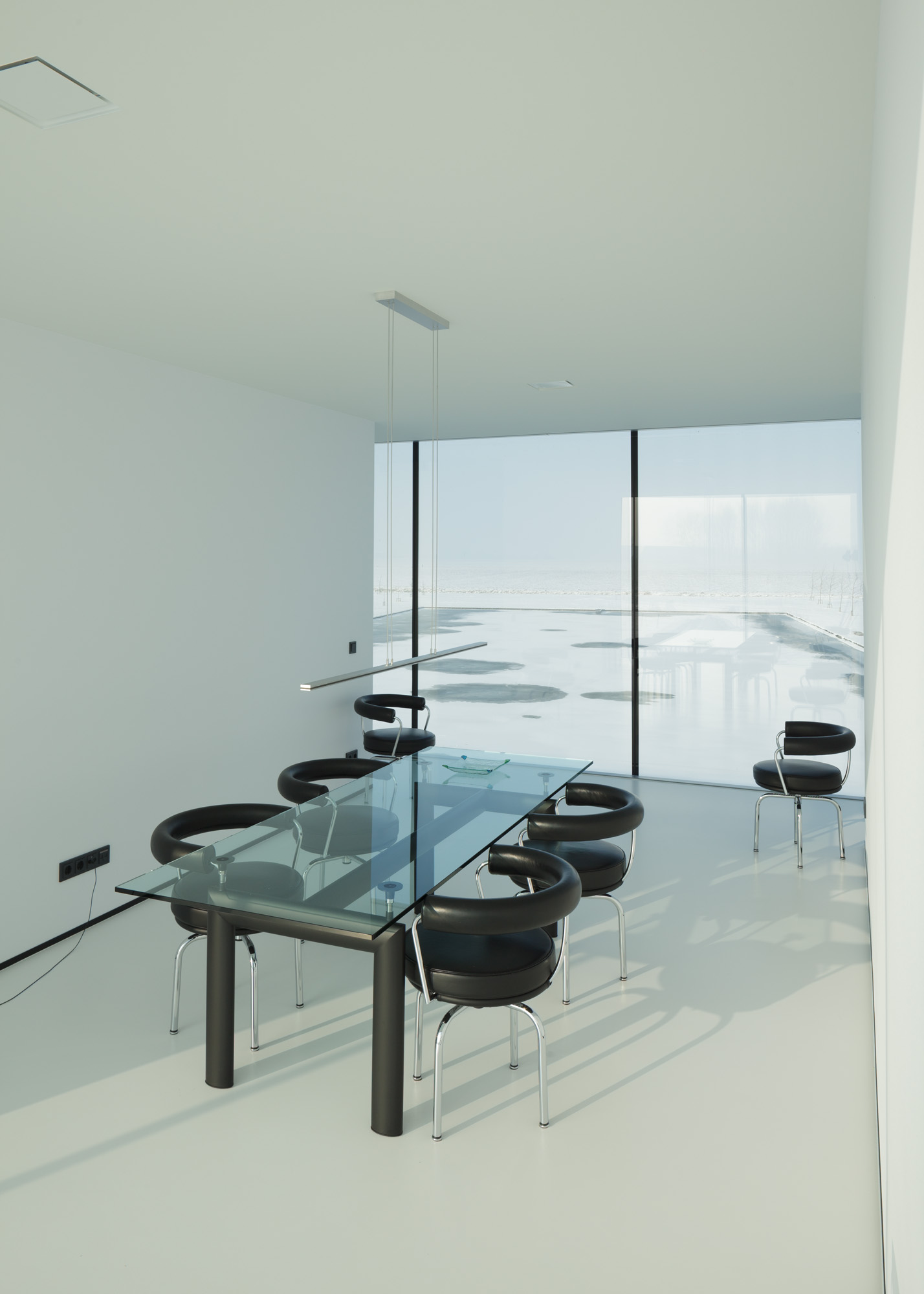 The dining room is a white, bare-walled room with a glass table and black semi-circular chairs. The floor-to-ceiling windows show the view of a bare, flat, snowy landscape.