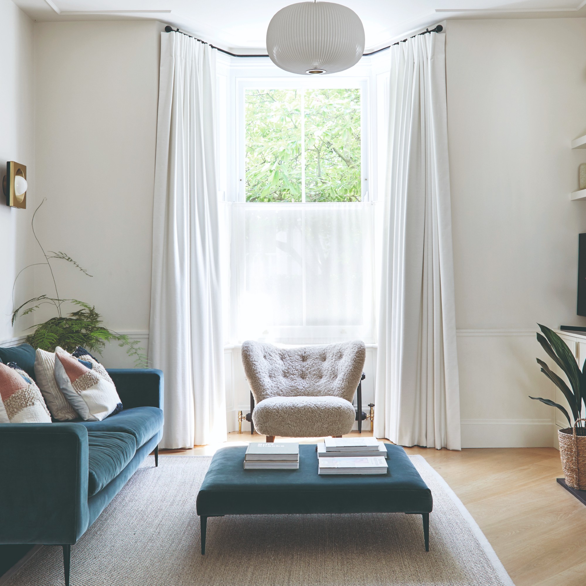 A light living room with a high ceiling and a bay window with floor-length pleated white curtains