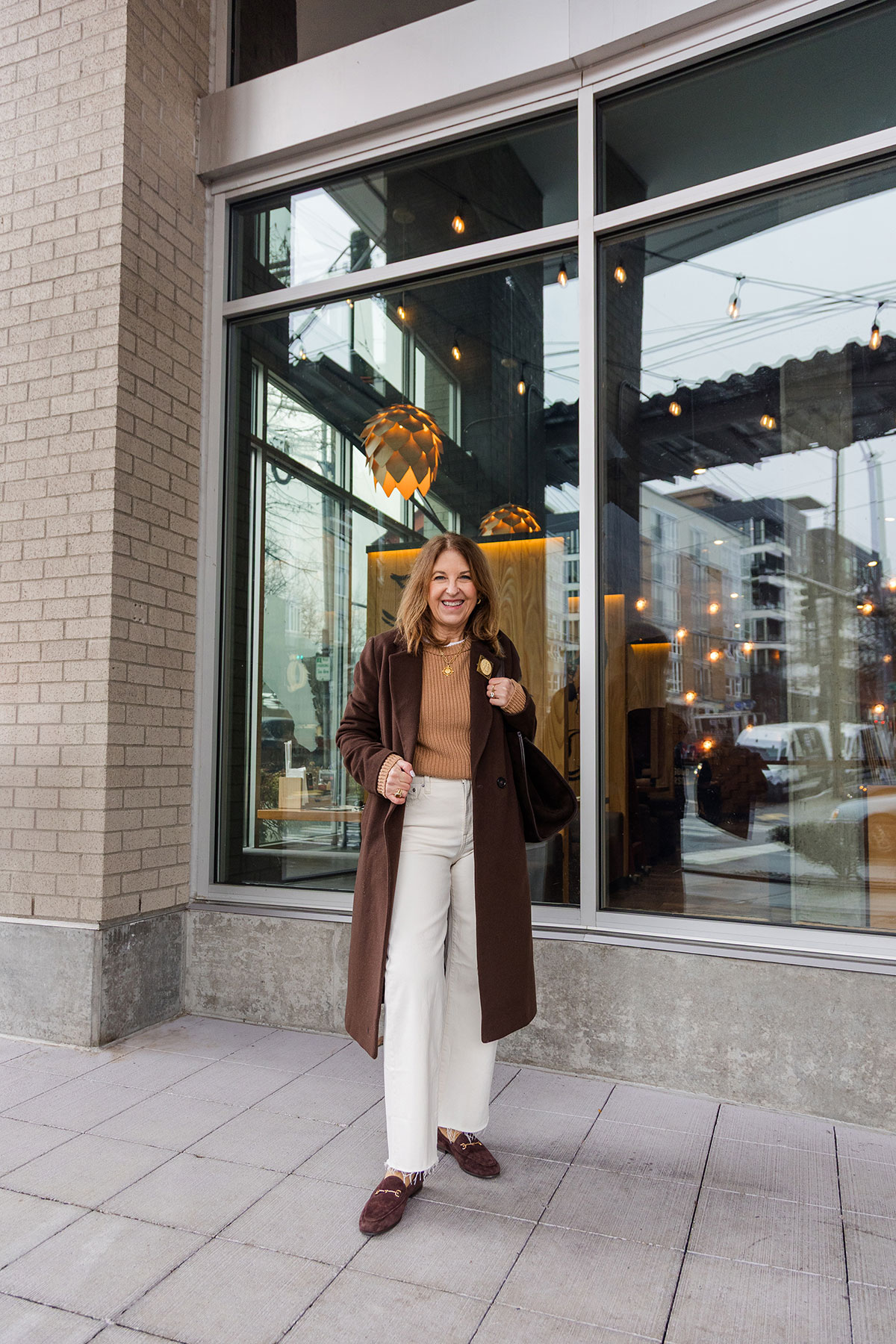 A former Nordstrom stylist wearing white jeans with a light brown sweater, a long dark brown coat, and dark brown loafers.