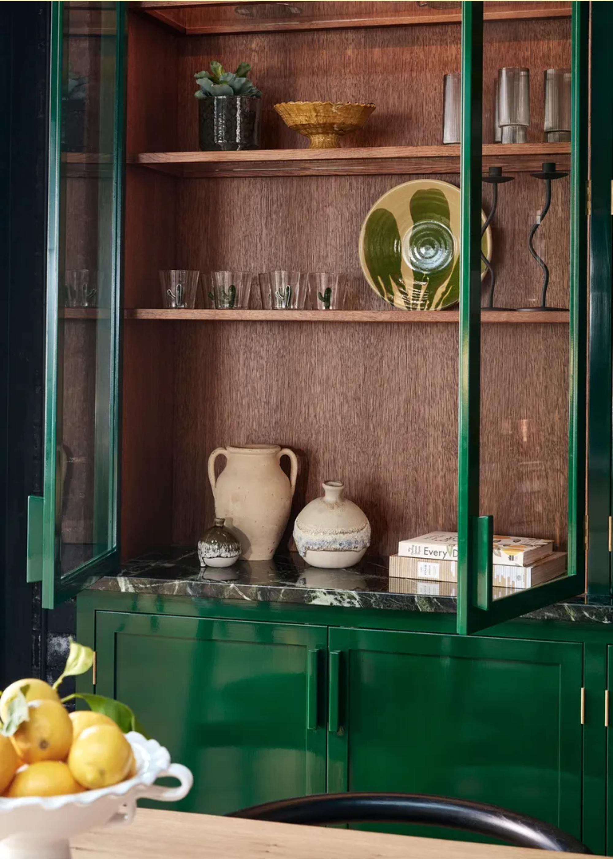 A glossy green cabinet with a ceramic plate, vases, glassware, and a potted plant by wavy candle holders
