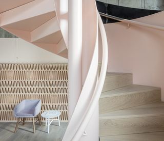 Spiral staircase in the Leman Locke hotel. Geometrically placed wooden boards in the shape of a triangle are covering the walls behind the staircase, with a lavender-colored suede chair, and a white table next to it. To the right and closer to us, the spiral staircase is painted in pale pink, with wooden stairs.