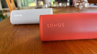 Sonos Roam in white behind Sonos Roam 2 in orange red on wooden desk