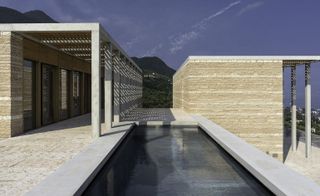 The exterior view of the Villa Eden. It uses timber, concrete structure, and local stone in sand color. We see a pool and mountains in the distance.