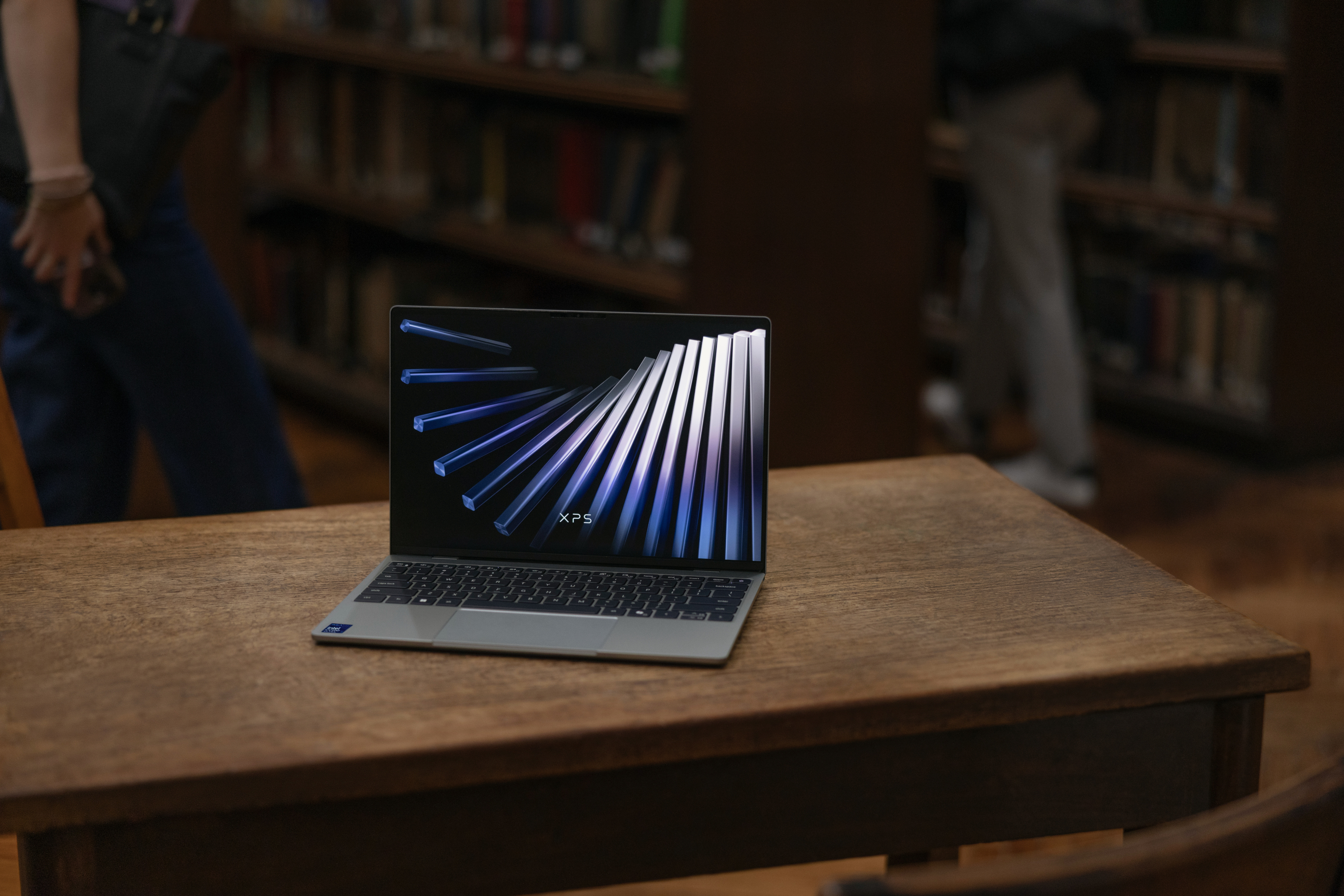 An XPS 13 laptop sitting open on a desk, with bookshelves in view behind it.