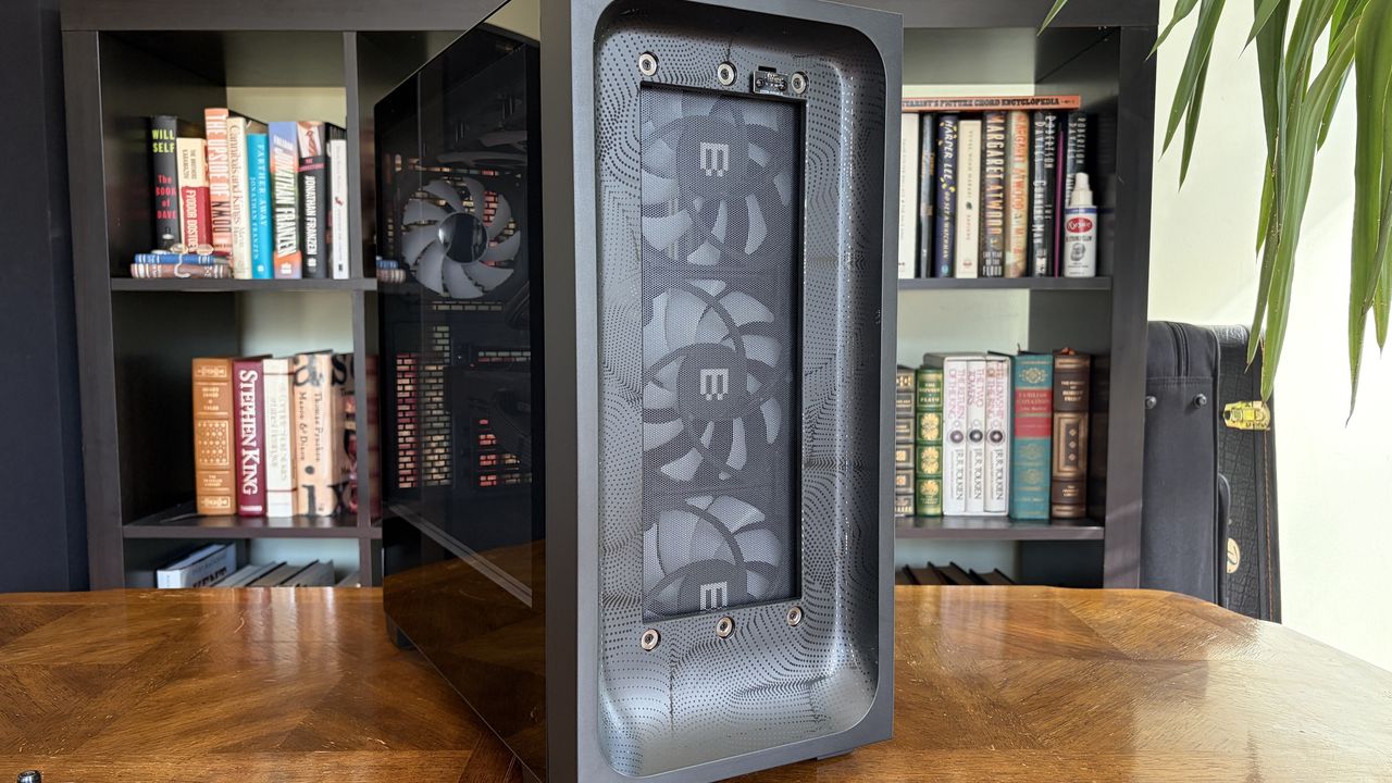 A sleek MG-1 MK.II gaming computer case with visible cooling fans sits on a wooden table. In the background, a bookshelf is filled with various books, adding a cozy atmosphere.