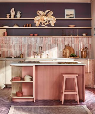 A kitchen with a curved dusky pink kitchen island with a white countertop, a brown flower pendant light above it, and dark purple walls with a light pink tiled splashback behind it