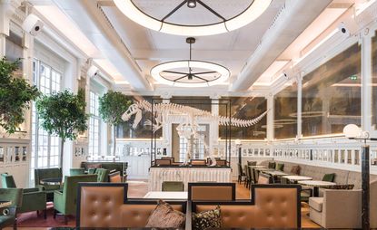 Interior look of the JNĉQUOI luxury restaurant. Tall ceilings and windows, which are in the french style. Various sitting arrangements, with the sitting area in beige, orange, and deep green, with white marble tables. At the center, there is a dinosaur&#039;s skeleton.