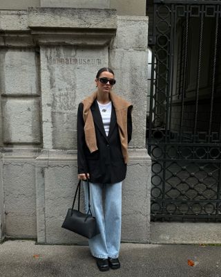 Mary wears a brown sweater, black blazer, jeans, white tank top, black handbag, and black loafers.