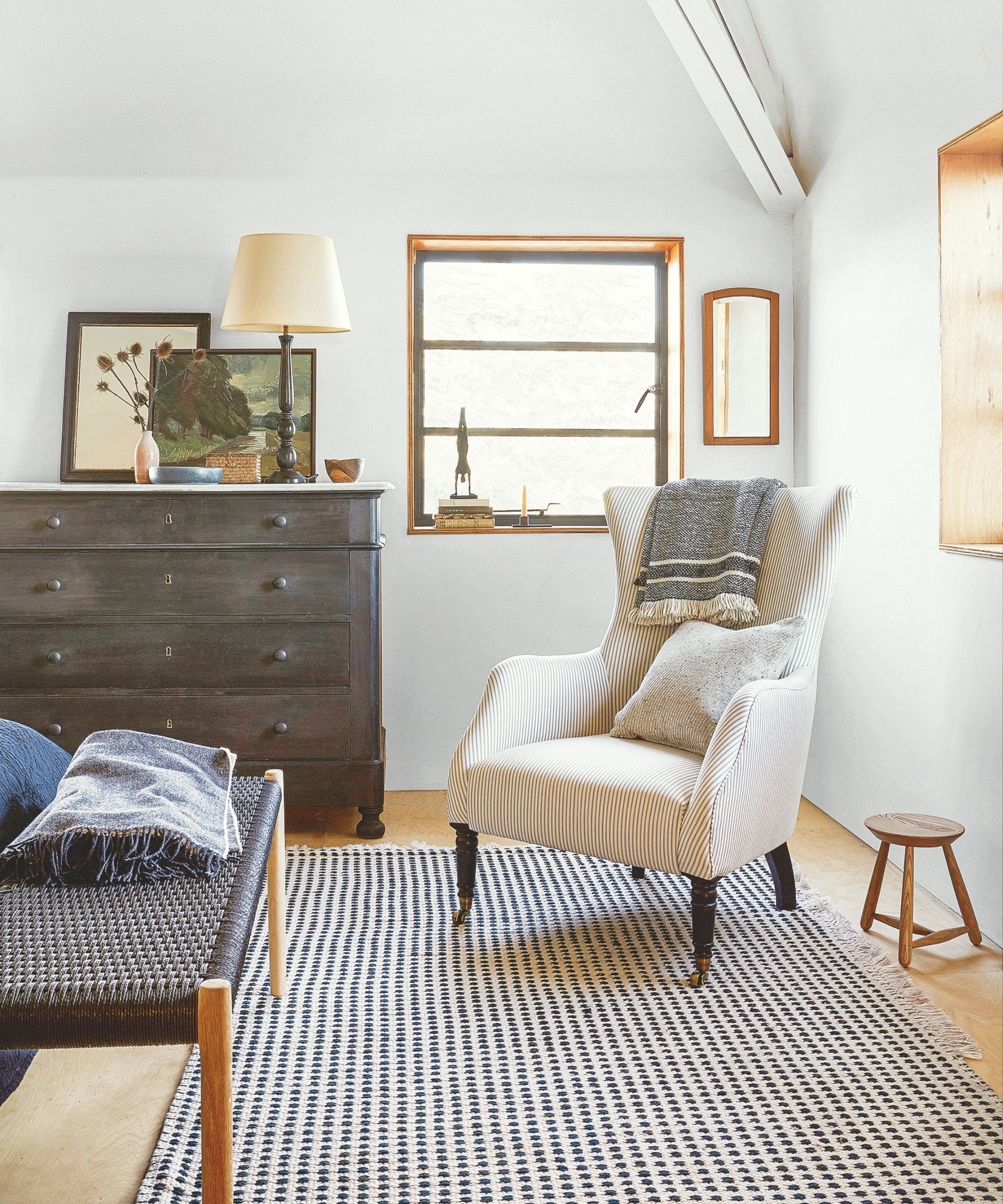 White bedroom with striped rug, white armchair, navy bench with wooden legs, and dark brown chest of drawers beside a large window.
