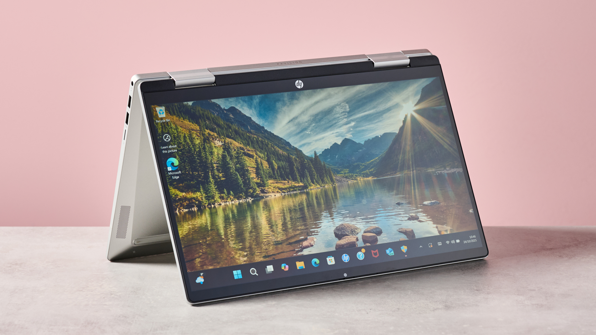 Three-quarter view of HP Pavilion x360 14 open on desk, flipped upside down in touchscreen mode, with pink wall in background