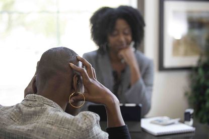 Two women talking over a desk with the woman in the foreground having her hands on her head in frustration