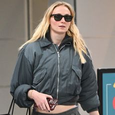 Sophie Turner arriving at JFK Airport wearing a gray bomber jacket, sweatpants, and Adidas blue and white Samba sneakers.