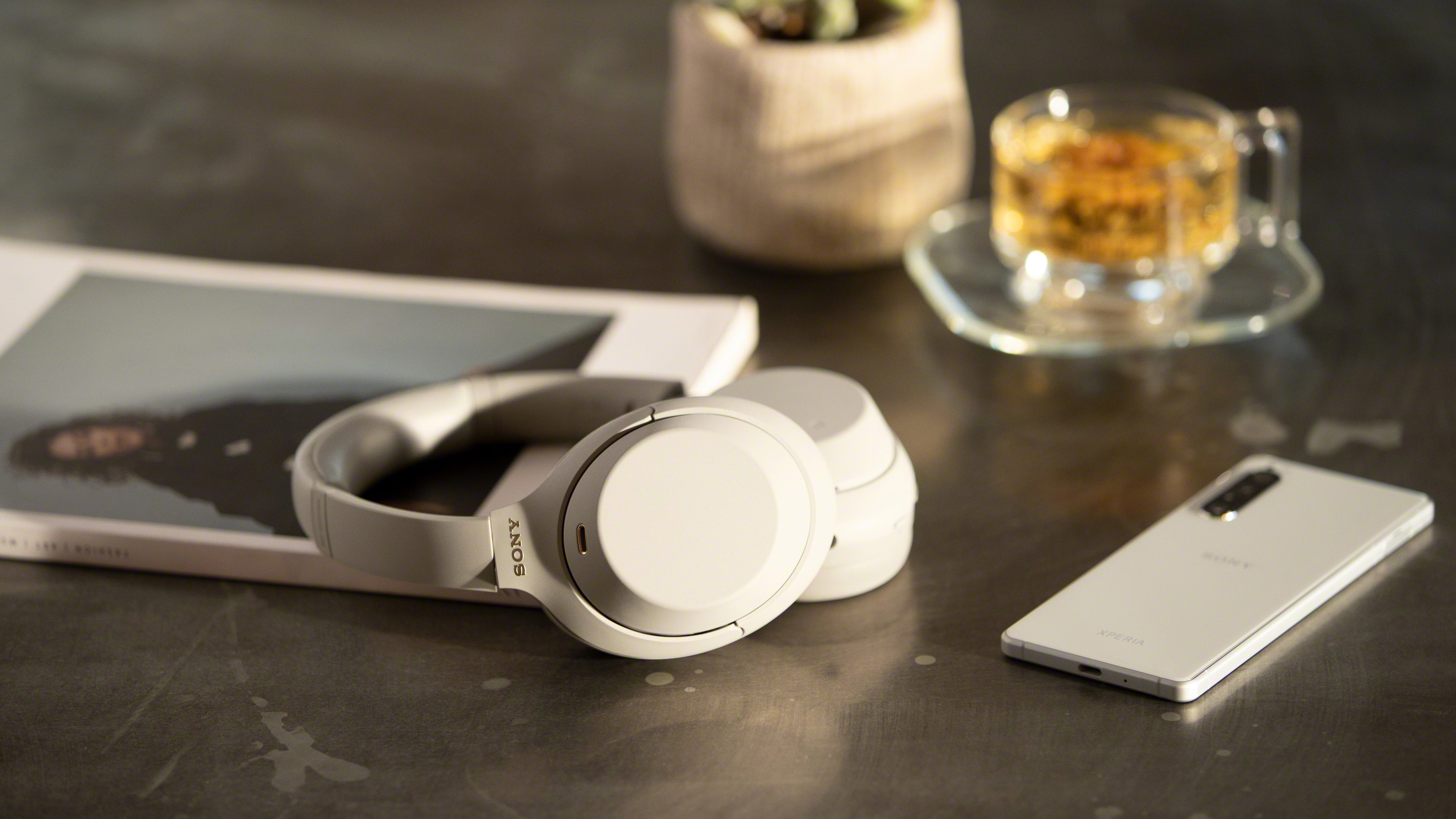 Sepasang headphone sony wh-1000xm4 berwarna putih di atas meja gelap di samping majalah dan smartphone berwarna perak