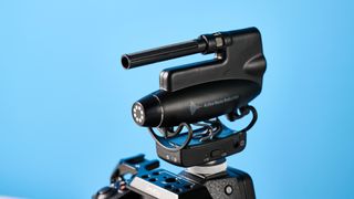 a black shotgun microphone by joby called the wavo pro is photographed by itself against a blue background, showing its muff, dead cat, attachment foot, and connection ports