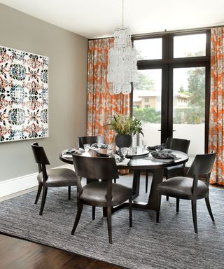 Gray dining room with black wooden dining table and chairs, orange patterned curtains, colorful artwork, chandelier hanging over table