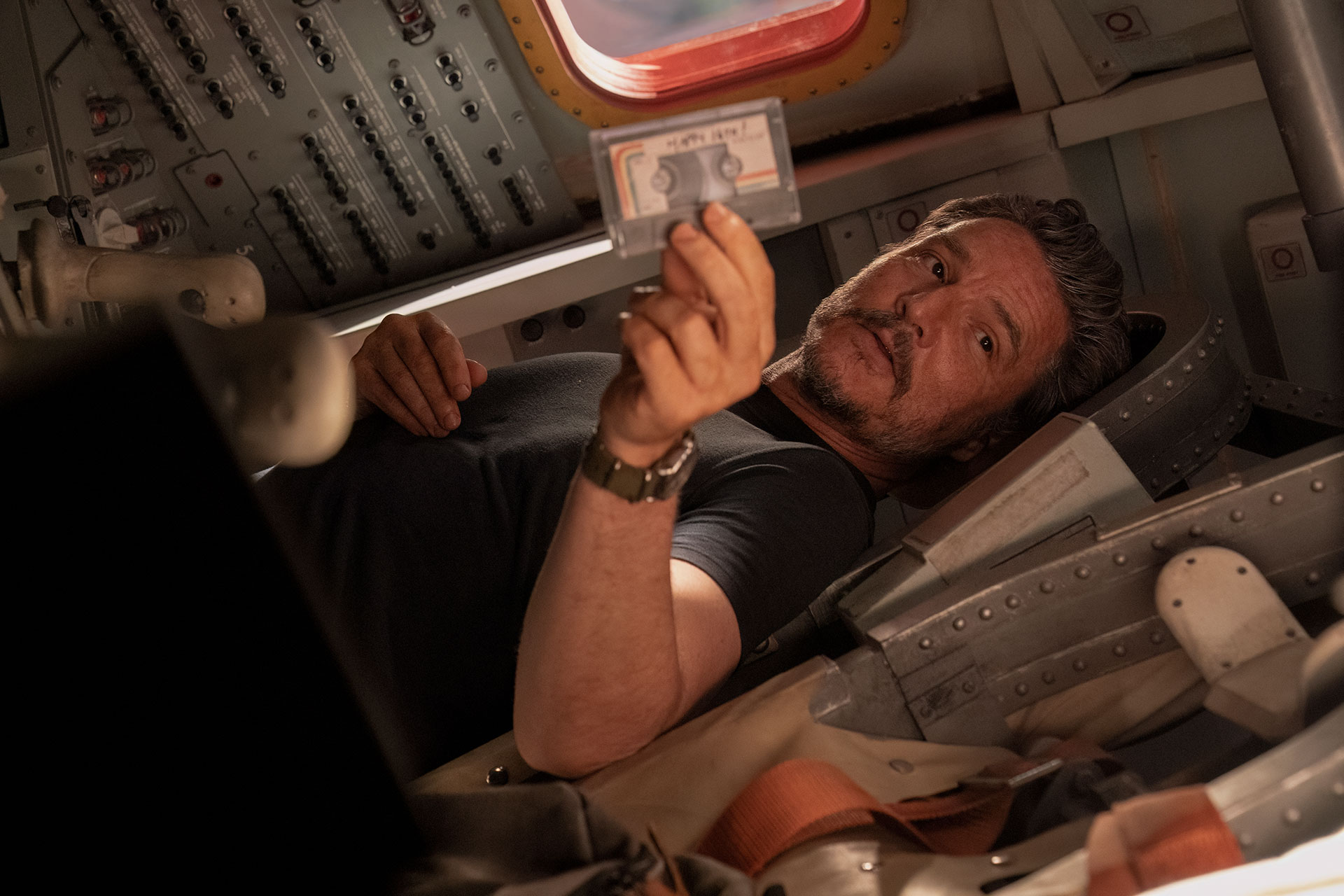 a man holds up a cassette tape while sitting inside a 1960s space capsule