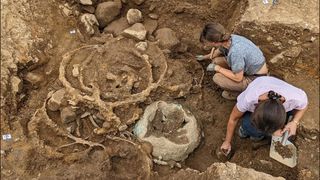 A look down into an excavation site, where two women crouch to the right, collecting samples from half-unearthed stones