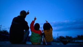 A man and a woman with a child between them all point at a night sky above them with a telescope on a tripod behind them